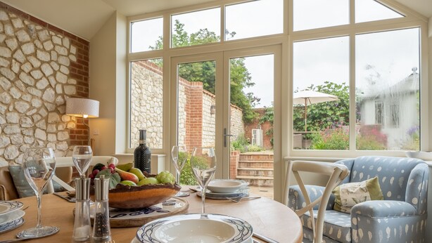 A dining area with a stone wall, a table set with plates, glasses, and a fruit bowl. There is a blue patterned armchair and a view of a garden through large windows. - Sutton Cottage, Thornham, Norfolk (Thornham)