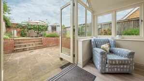A room with a large sliding glass door leading to a patio with a table and chairs, a blue patterned armchair, and a view of a brick wall and garden. - Sutton Cottage, Thornham, Norfolk (Thornham)