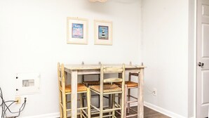 Une pièce avec un mur blanc, une table en bois avec des chaises, deux affiches encadrées, un luminaire suspendu et une porte.