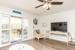 A room with a ceiling fan, a TV, a clock, a chair, and a sign that reads 'Let's do Sunset'. - Aruba Sun - 1 BR Surf Condo - Great Views and Community Pool (Surf City)
