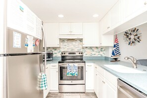 A kitchen with white cabinets, a stainless steel refrigerator, a microwave, an oven, and a sink with a decorative sailboat. - Aruba Sun - 1 BR Surf Condo - Great Views and Community Pool (Surf City)