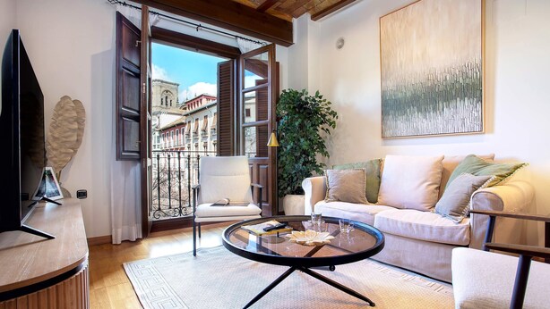 Ein Wohnzimmer mit rundem Couchtisch, einer Couch, einem Fernseher und einem Balkon mit Blick auf Gebäude.