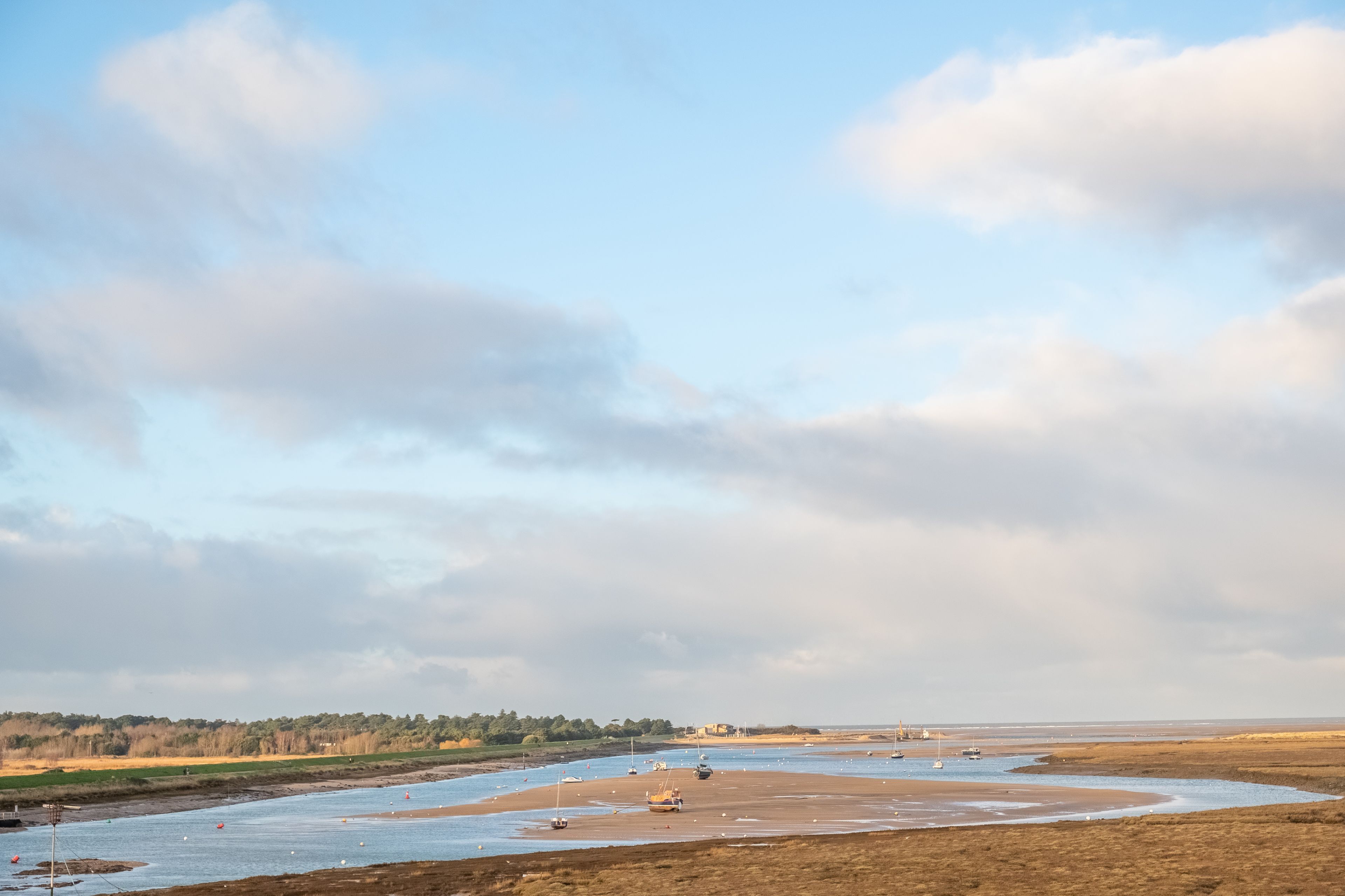 A coastal landscape with boats on a wide, shallow water body, surrounded by flat terrain and distant trees.