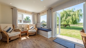 A spacious living area with a large window, wicker furniture, and a view of a garden. - Tanholt, Holme-next-the-Sea, Norfolk (Holme-next-the-Sea)