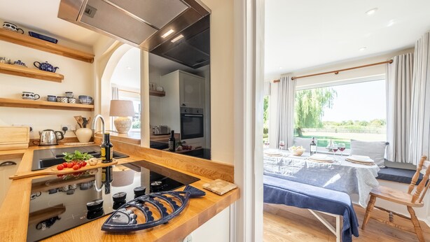 A modern kitchen with wooden shelves, a built-in stove, and a dining area with a table and chairs. - Tanholt, Holme-next-the-Sea, Norfolk (Holme-next-the-Sea)