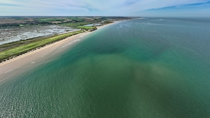 Ovenfra set kystlandskab med sandstrand, grønne marker og et stort vandareal.
