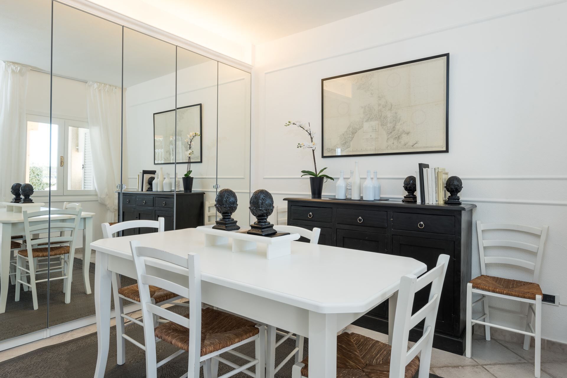 A dining area with a white table and chairs, a black cabinet with decorative items, and a large mirror reflecting the room.