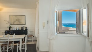 A room with a dining area, a view of the sea, and a window with white curtains. - Villaggio S'abba e Sa Pedra - S'ABBA 22 (07020 Golfo Aranci OT)