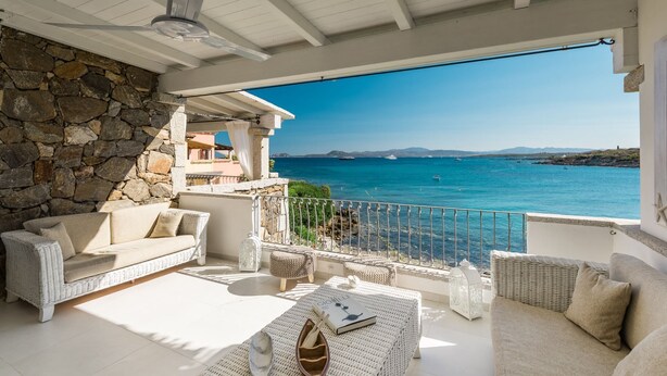 Une terrasse ensoleillée, meublée de mobilier en osier, avec un mur en pierre et une vue sur la mer.