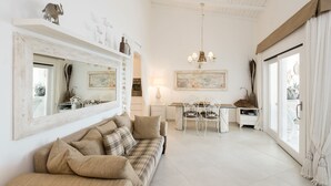 Un salon spacieux avec un grand canapé, une table à manger et des chaises. On y trouve un lustre, des œuvres d’art accrochées aux murs et une vue sur l’extérieur grâce à une grande fenêtre.