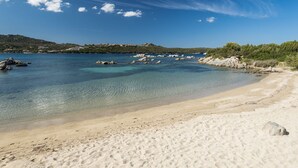 Une plage de sable fin bordée d’une eau claire, ponctuée de formations rocheuses et offrant une vue sur une colline lointaine où se dressent des bâtiments.