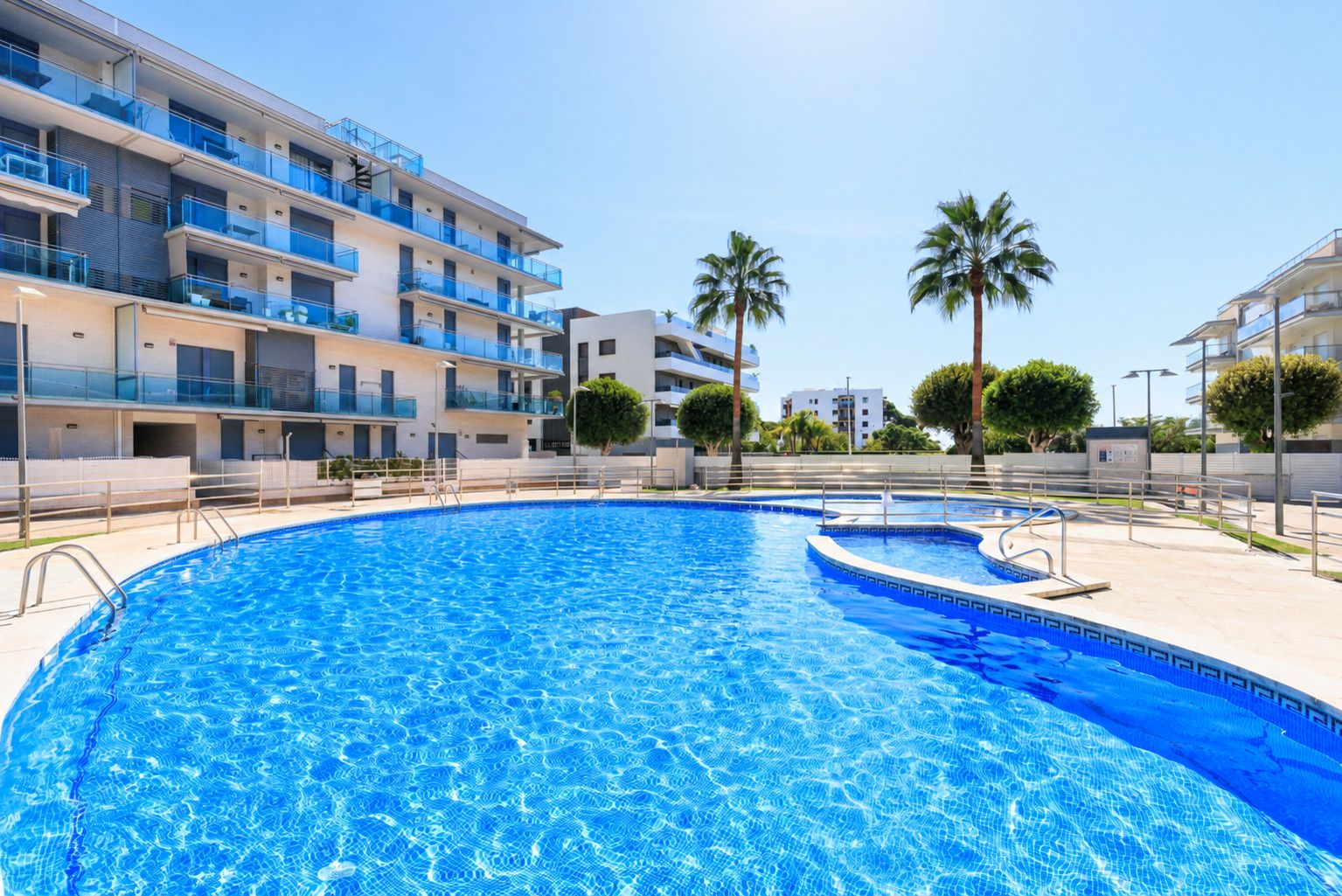 Une piscine entourée de bâtiments modernes dotés de balcons, de palmiers et surplombant un ciel dégagé.