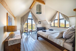 A bedroom with a large bed, wooden floor, a desk with a lamp, and a view of the outside.