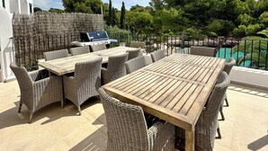 En uteplats med ett träbord och flätade stolar, en grill och en pool i bakgrunden.