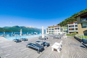Una terraza de madera con tumbonas y sombrillas, una piscina y un edificio con balcones con vistas a un paisaje montañoso.
