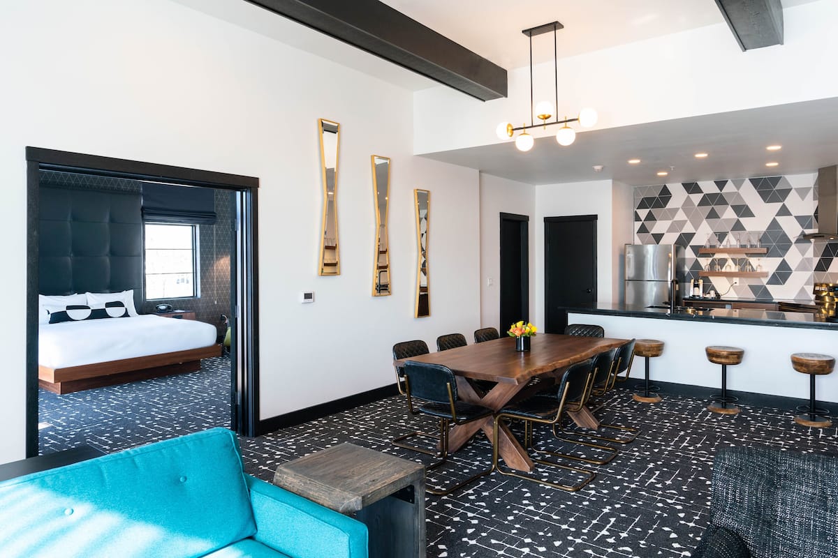 A modern hotel room with a large bed, a dining area with a wooden table and chairs, and a kitchenette.