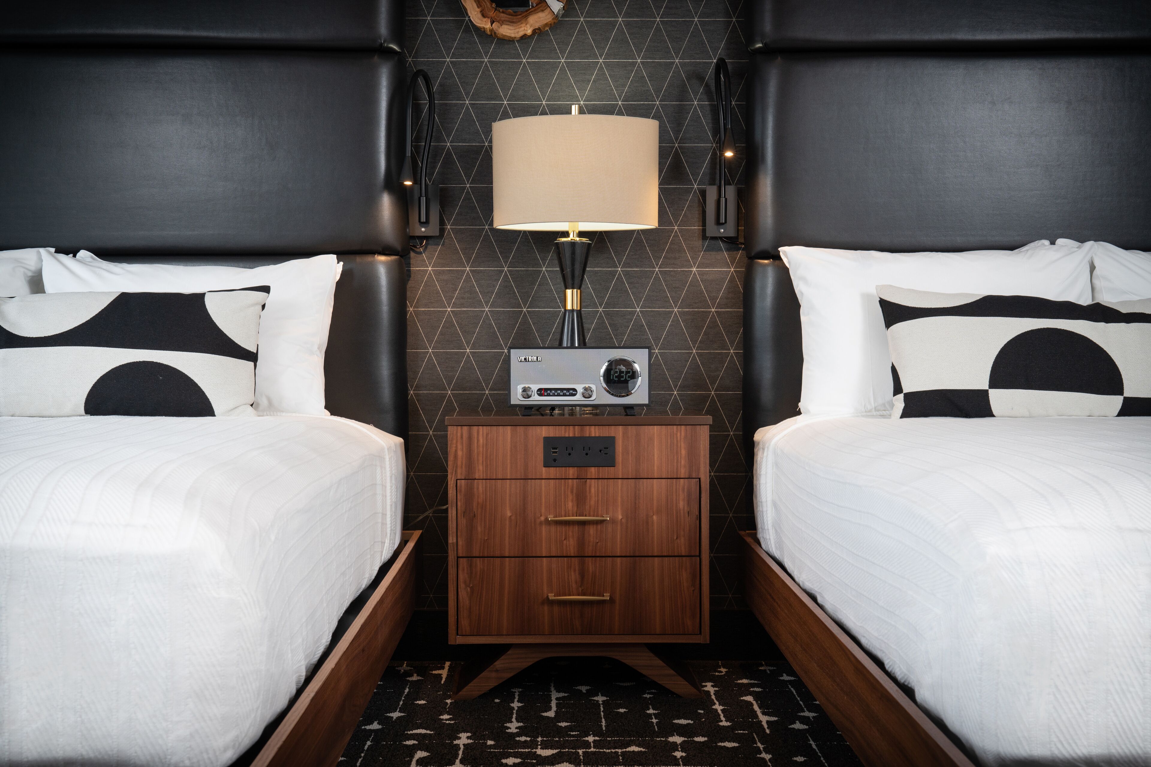 A hotel room with two beds, a nightstand with a radio, and a wall-mounted lamp.