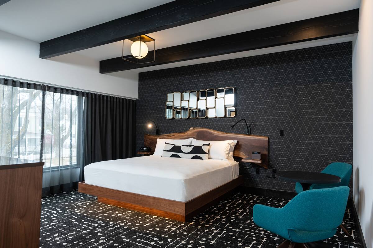 A modern hotel room with a large bed, a wooden headboard, a black tiled wall, a round table, and a teal chair.