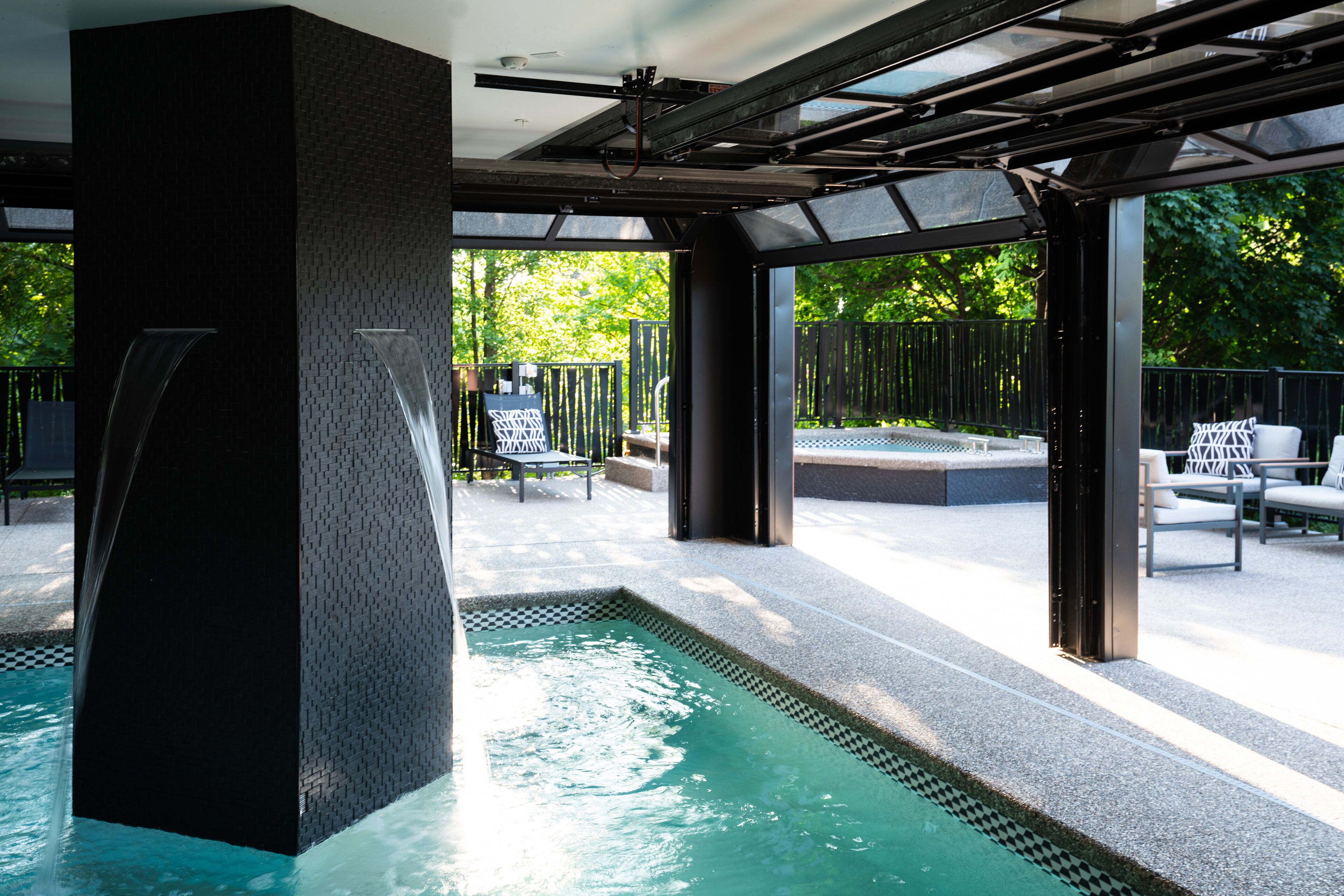 A modern outdoor pool area with a geometric water feature, seating, and a view of greenery.