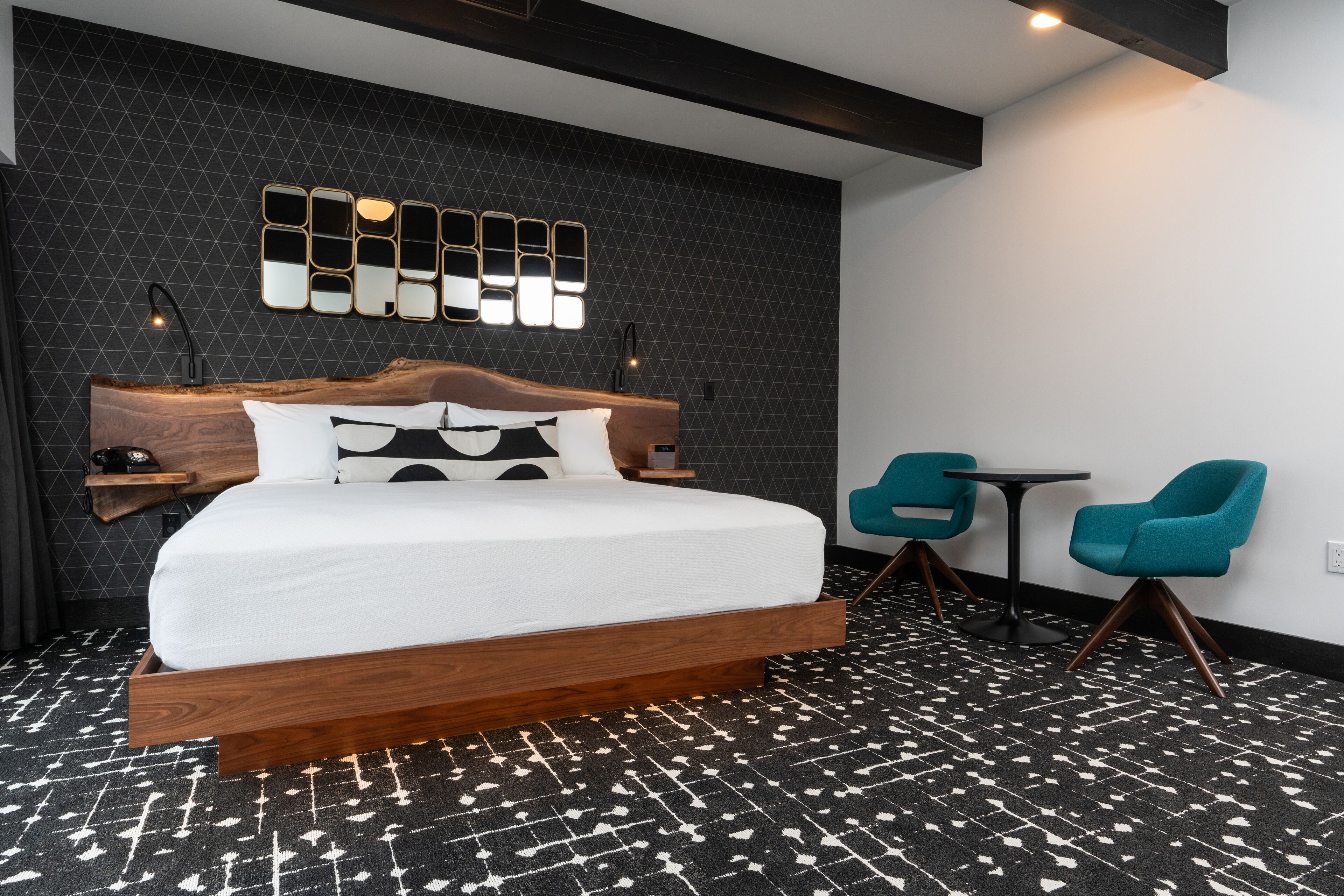 A modern hotel room with a large bed, a wooden headboard, a wall-mounted mirror, a small table, two chairs, and a patterned carpet.