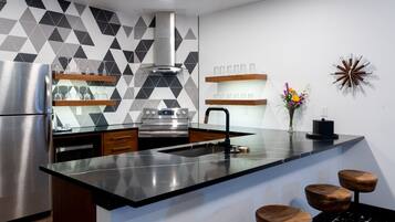 Una cocina moderna con un refrigerador de acero inoxidable, horno empotrado y encimera negra. Hay taburetes de madera y un elemento decorativo en la pared.