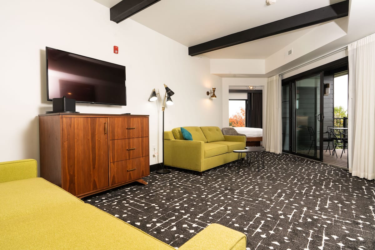 A modern hotel room with a flat-screen TV, wooden cabinet, yellow sofa, green sofa, and a patterned carpet.
