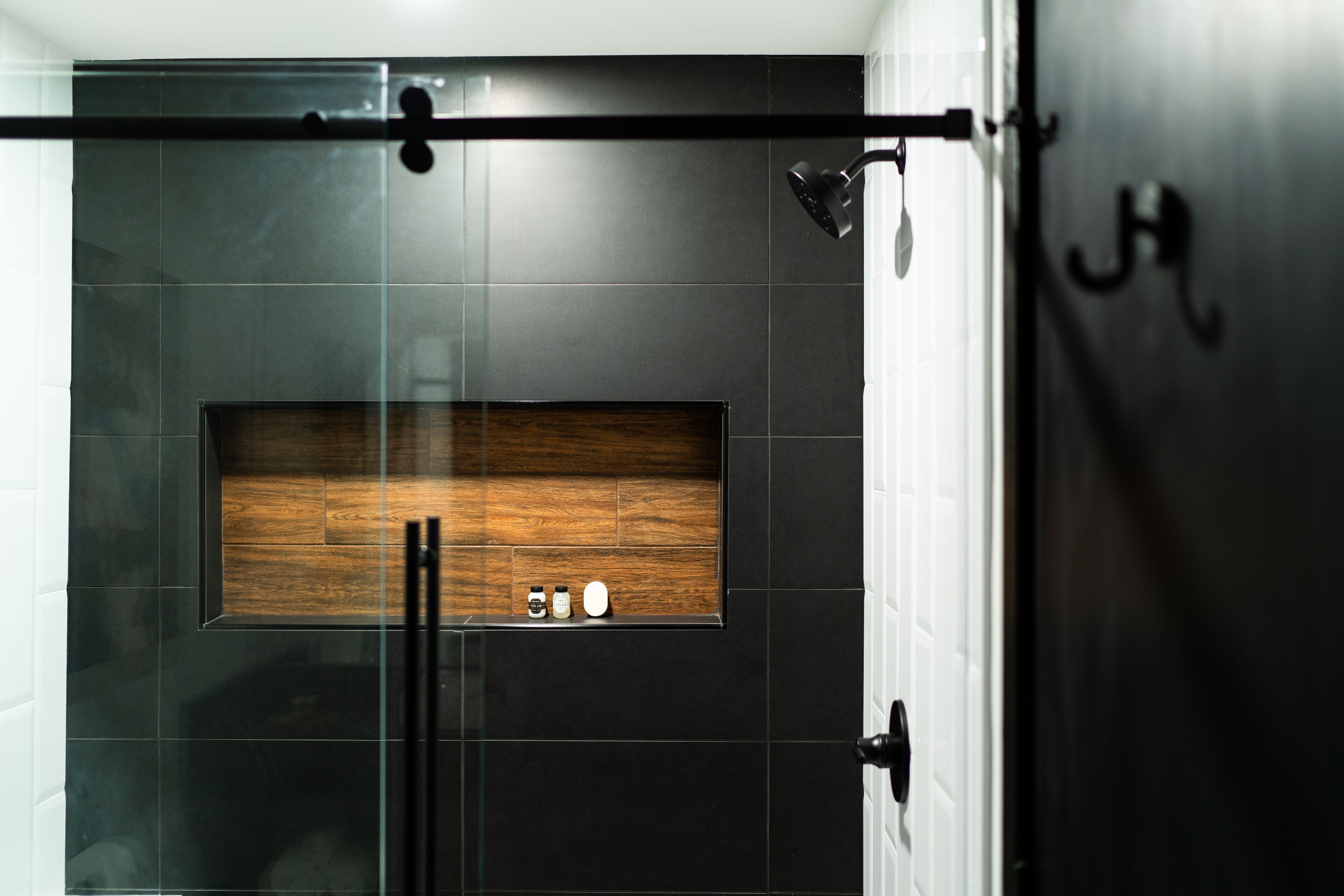 A modern bathroom with a glass shower enclosure, a wooden shelf with toiletries, and a black door with a handle.