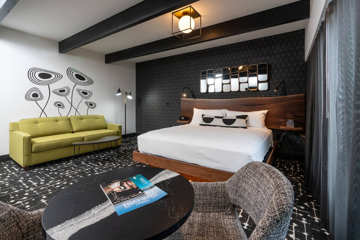 A modern hotel room with a bed, a green sofa, a round coffee table with a magazine, and a unique wall art piece.