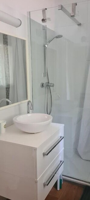 Moderni kylpyhuone, jossa on lasiseinällä varustettu suihku, valkoinen pesuallas ja peili, joka on sijoitettu pesualtaan yläpuolelle.