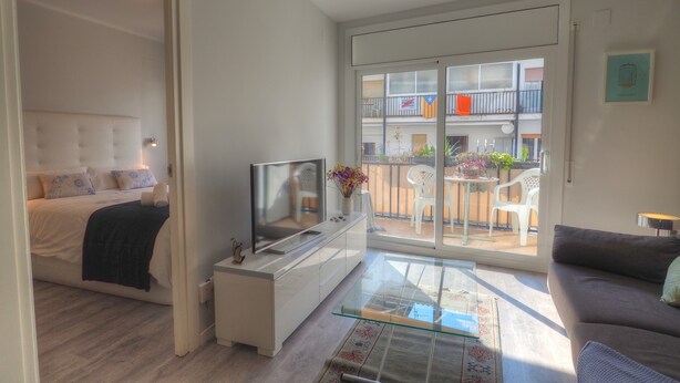 A modern living room with a bed, a TV, a glass coffee table, and a balcony with outdoor furniture.