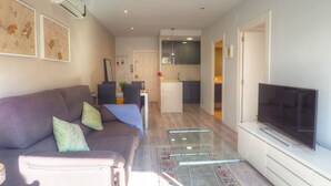 A living room with a sofa, a glass coffee table, a flat-screen TV, and a kitchen in the background.