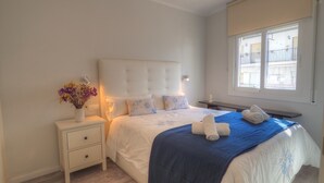 A bedroom with a large bed, a bedside table with a vase of flowers, and a window with a view of buildings.