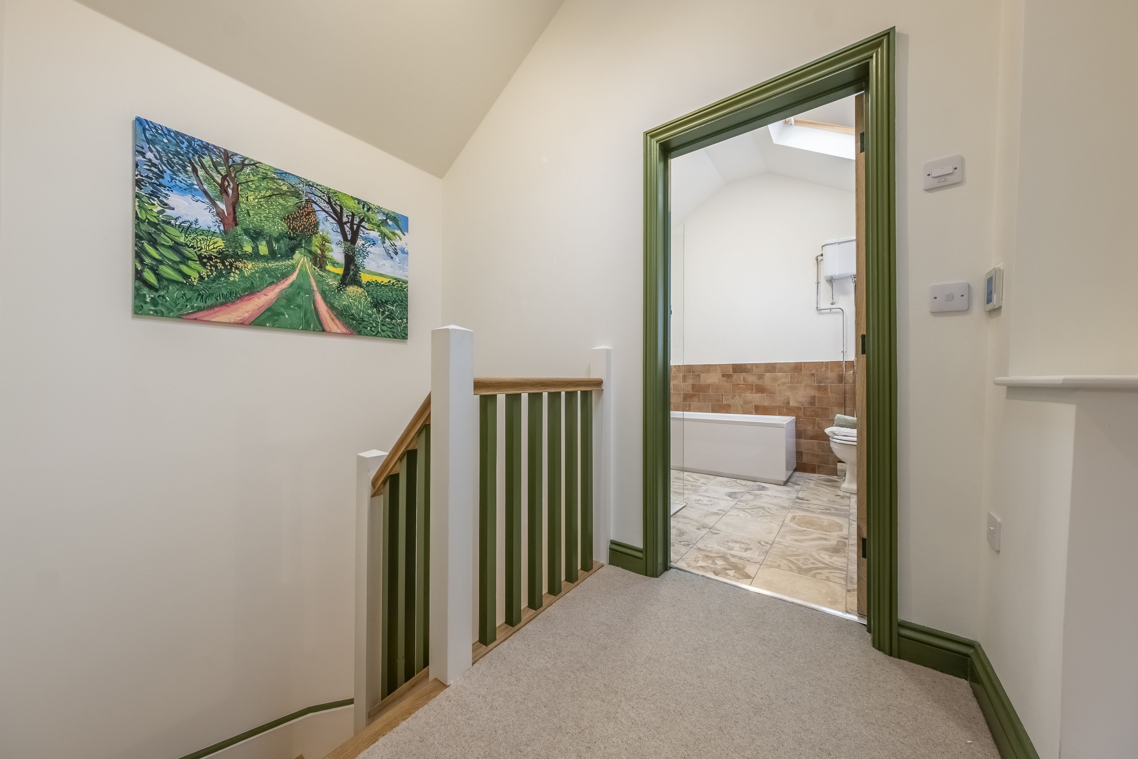 A hallway with a painting of a landscape, a wooden railing, and a view into a bathroom with a bathtub and toilet.