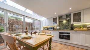 A modern kitchen with white cabinets, a wooden dining table, and a large window. - Mill House Cottage, Brancaster, Norfolk (King's Lynn)