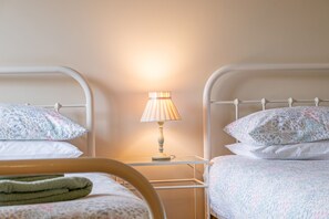 A room with two beds, each with floral-patterned bedding, a bedside table with a lamp, and a wooden chair.