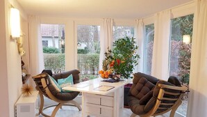 Ein Zimmer mit Holzboden, einem weißen Schreibtisch, zwei braunen Stühlen, einem kleinen weißen Heizgerät und einem Fenster mit Vorhängen.