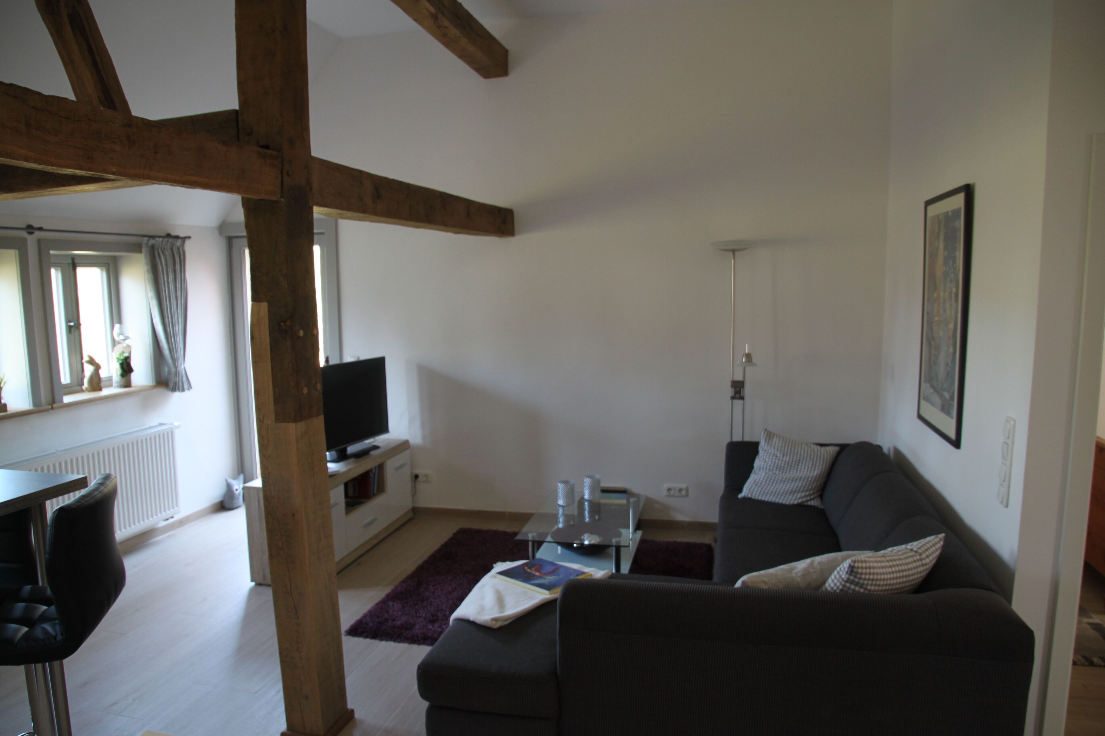 Ein Wohnzimmer mit einem dunklen Sofa, einem Glastisch und einem Fernseher. Ein Fenster mit Vorhängen und eine Heizung sind ebenfalls vorhanden.
