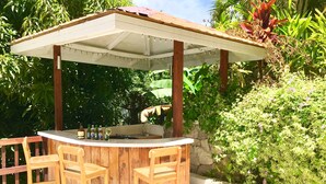 Area bar esterna con tavolo in legno e due sedie, bancone in pietra e una pergola sovrastante.