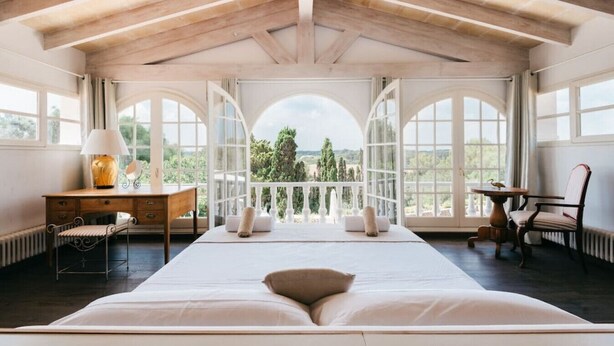 Un'ampia camera da letto con un letto grande, una scrivania con lampada, una sedia e vista all'aperto attraverso ampie finestre.