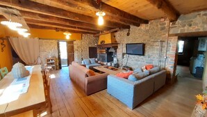 Un intérieur rustique avec des murs en pierre, des poutres en bois, une cheminée en pierre, un téléviseur et un coin repas comprenant une table et des chaises.