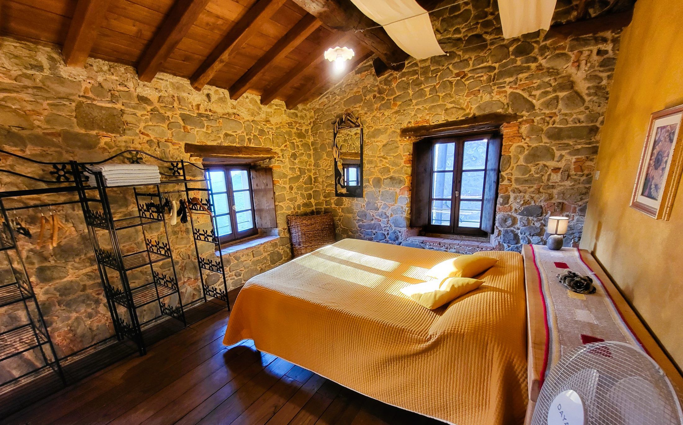Ein Schlafzimmer mit Steinwand, Holzbalken, einem Bett mit gelber Decke, einem Ventilator und einem Fenster mit Fensterläden.