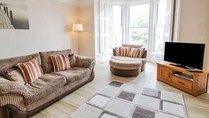 A living room with a sofa, ottoman, TV stand, and large windows. - Ayr Beach View Apartment (Ayr)