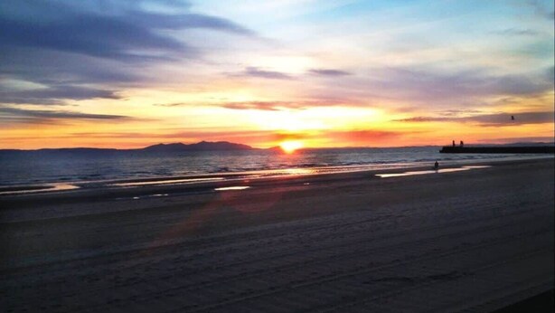 A beach with a sunset, calm sea, and distant mountains. - Ayr Beach View Apartment (Ayr)