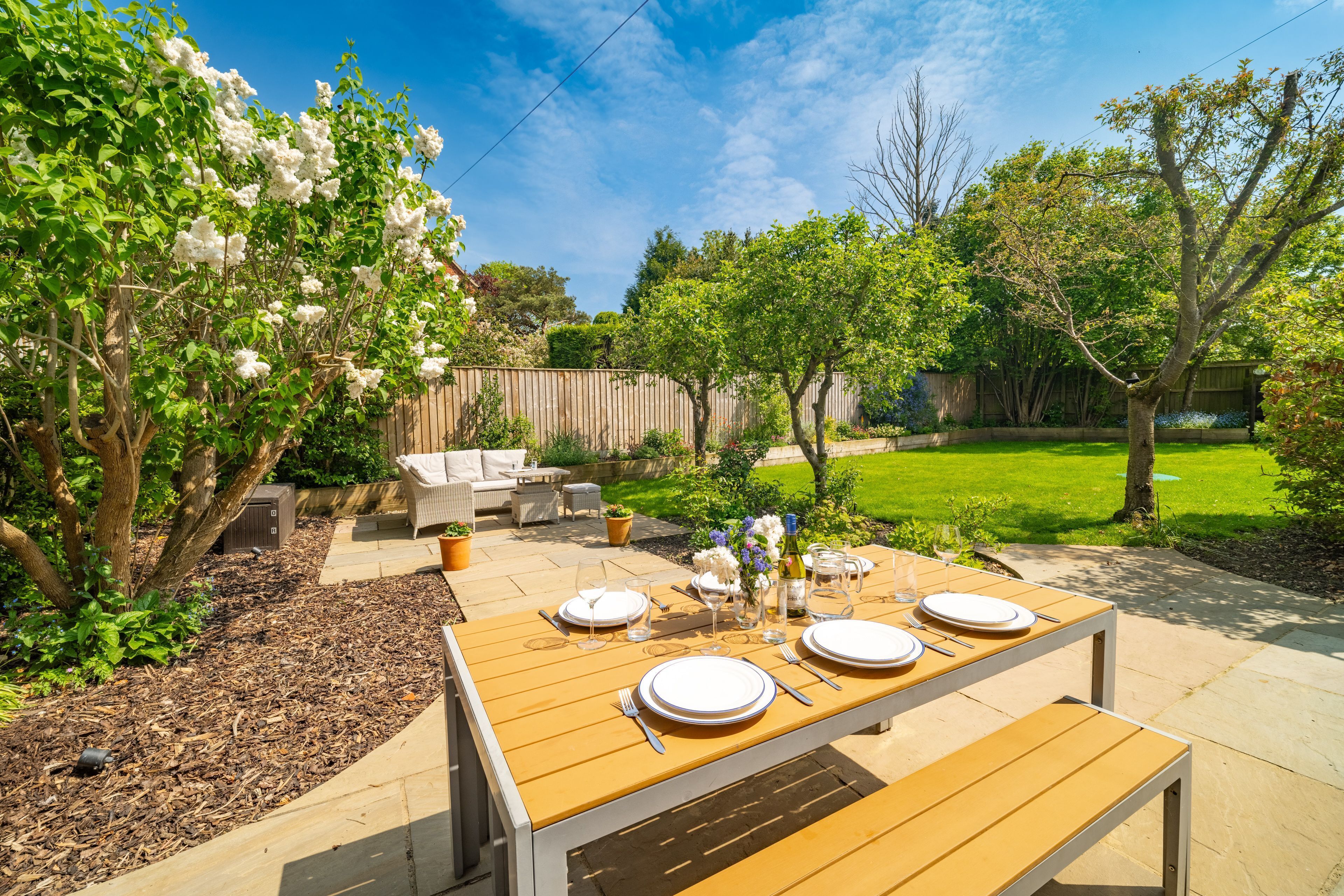 An outdoor patio with a wooden table set for four, surrounded by lush greenery and a wooden fence.