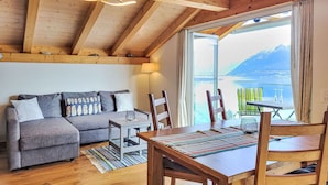 A cozy living room with a wooden ceiling, a sofa with patterned cushions, a dining table with chairs, and a large window offering a scenic view. - Beautiful apartment for 4 people with WIFI, TV, balcony, pets allowed and panoramic view (Porto Ronco)