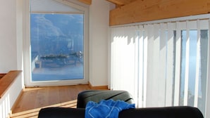 A room with a large window offering a view of a lake and mountains, a wooden ceiling, and a black sofa with a blue throw. - Beautiful apartment for 4 people with WIFI, TV, balcony, pets allowed and panoramic view (Porto Ronco)