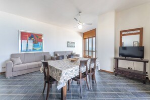 A dining area with a table, chairs, a ceiling fan, a television on a stand, and a sofa.