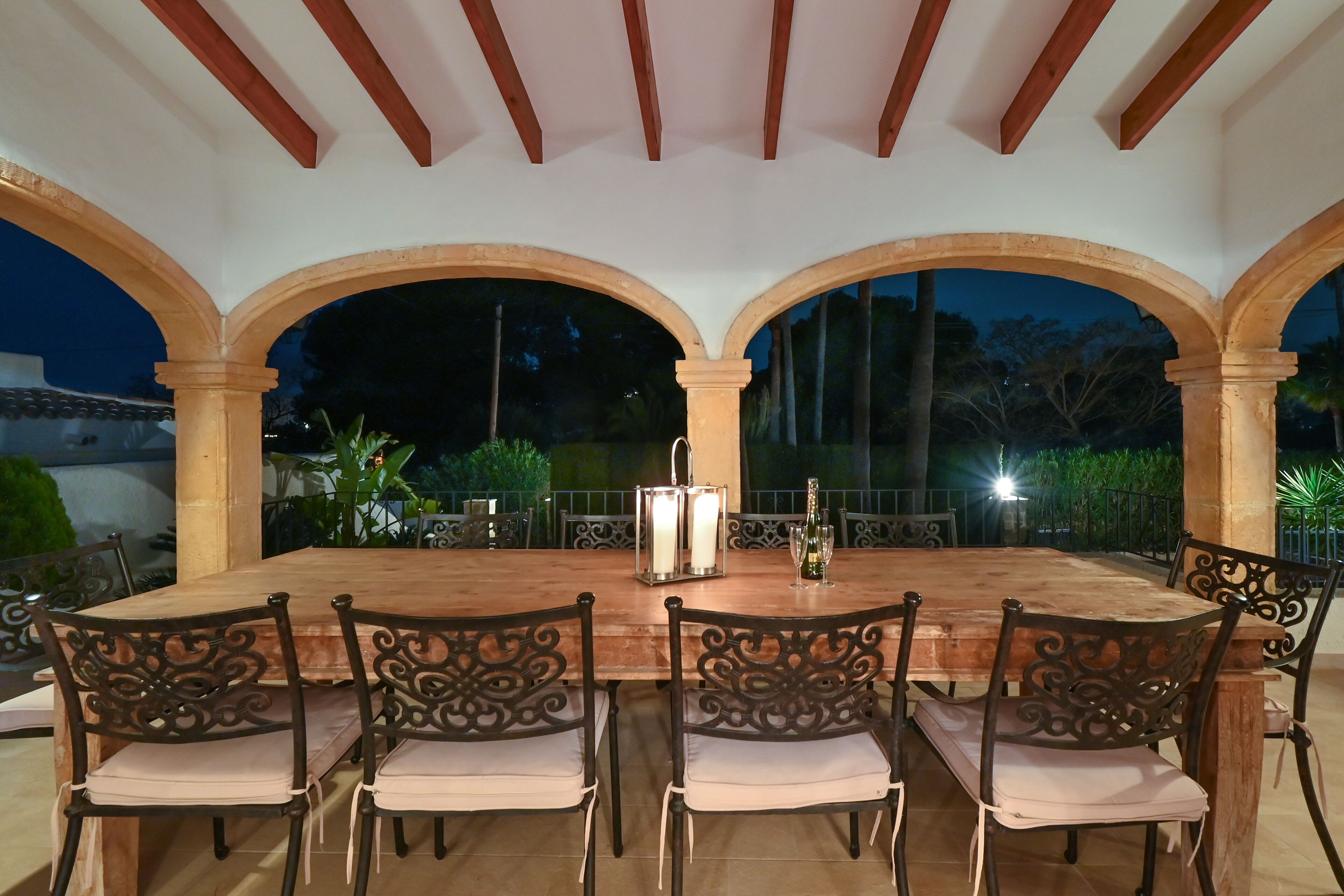 An outdoor dining area with a wooden table and chairs, surrounded by arches, and featuring a candle and a bottle on the table.