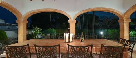 An outdoor dining area with a wooden table and chairs, surrounded by arches, and featuring a candle and a bottle on the table.