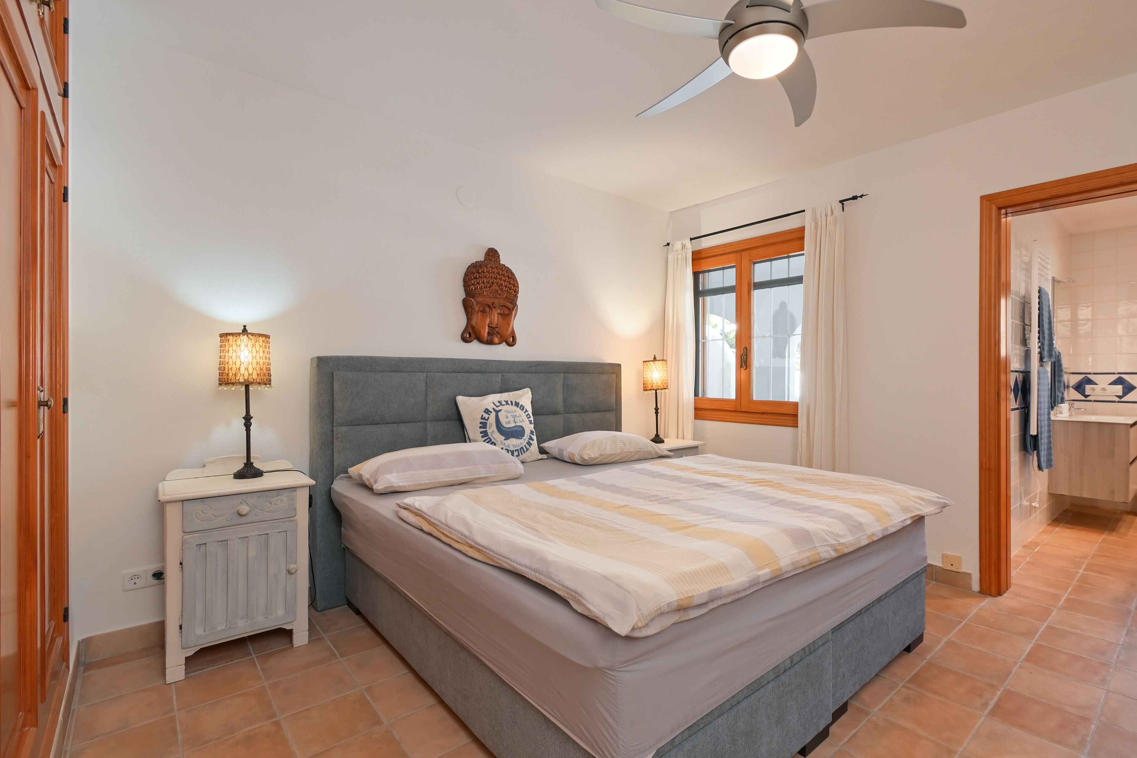 Un dormitorio con cama, mesitas de noche, ventilador de techo, ventana con cortinas y un baño visible a través de una puerta abierta.